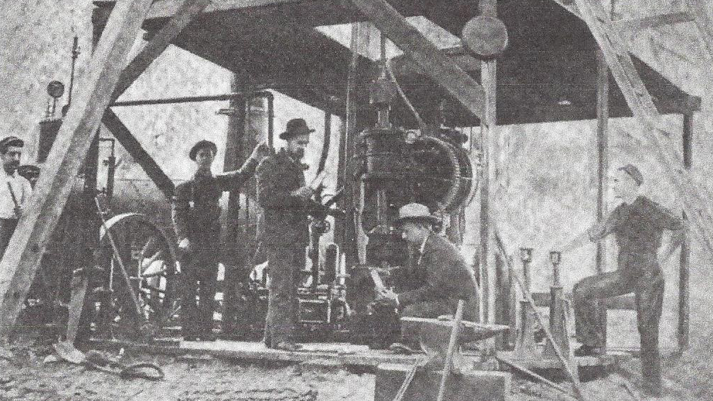 Black and white photo of several men operating and observing early industrial machinery under a wooden structure, wearing hats and work clothes, likely from the late 19th or early 20th century.