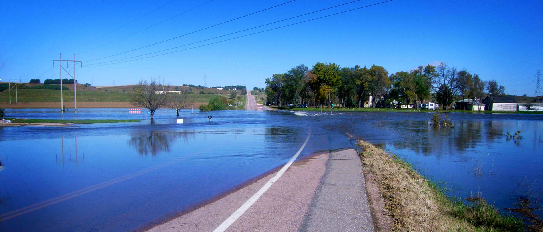 A rural road is flooded, with water covering both lanes and spreading into nearby fields and yards. Power lines run overhead, and trees and houses are seen in the background under a clear blue sky.