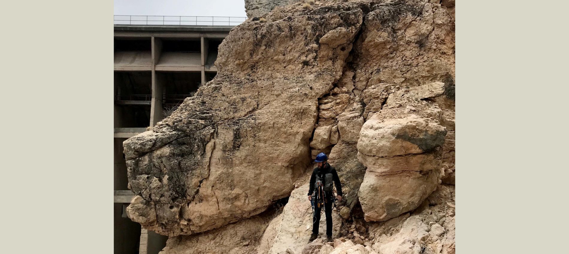 A rock climber wearing a blue helmet and climbing gear stands on a rugged rock face, with a large concrete structure visible in the background.