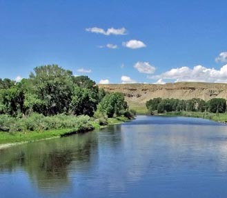 A calm river flows past green trees and grassy banks under a blue sky with a few white clouds. Low hills or bluffs are visible in the background.