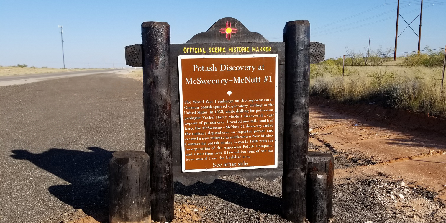 A brown historical marker sign on a roadside describes the potash discovery at McSweeney-McNutt #1, with a New Mexico state symbol on top and a dirt road and dry landscape in the background.