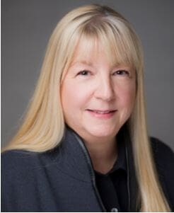 A woman with long straight blonde hair and bangs, wearing a dark jacket over a black top, smiles softly at the camera against a plain gray background.