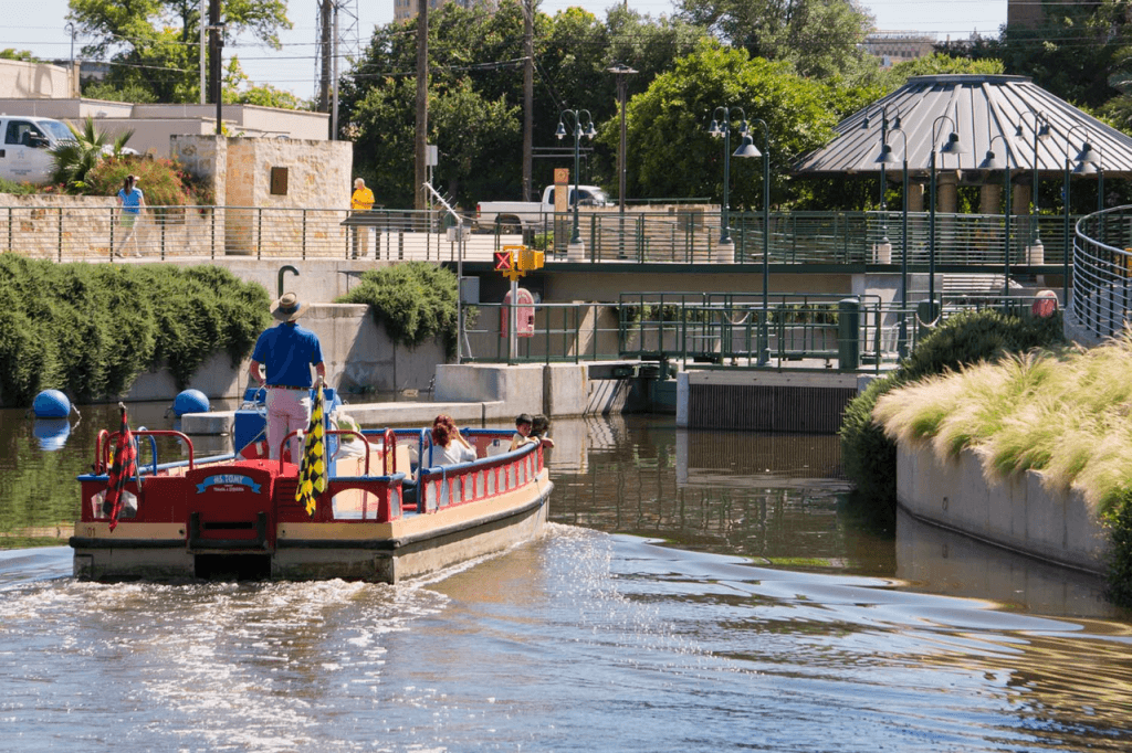 A small red riverboat with passengers is guided by a standing operator along a calm canal, passing greenery and heading toward a riverside pavilion on a sunny day.