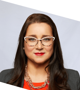 A woman with long brown hair wearing white-framed glasses, a gray blazer, a red top, and a statement necklace, poses in front of a light background. The image is slightly tilted.