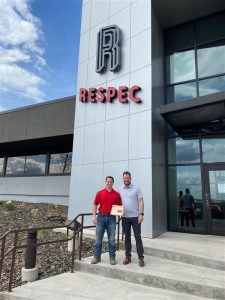 Two men stand smiling outside a modern building with large windows and a sign that reads RESPEC. One man holds a plaque. They are near the building’s entrance on concrete steps.
