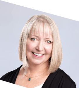 A smiling woman with straight, shoulder-length blonde hair and bangs, wearing a black top and a turquoise pendant necklace, posed against a light gray background. The image is slightly tilted.