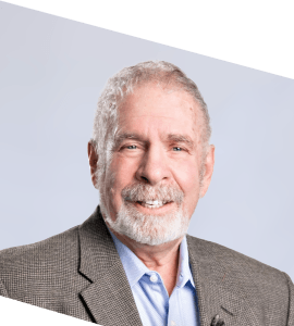 An older man with gray hair and a beard is smiling, wearing a gray blazer and blue shirt, posed against a light gray background. The image is slightly tilted.