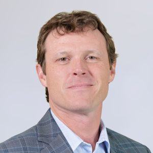 A man with short, wavy brown hair wearing a gray plaid suit jacket and light blue shirt, smiling slightly, with a neutral light gray background.