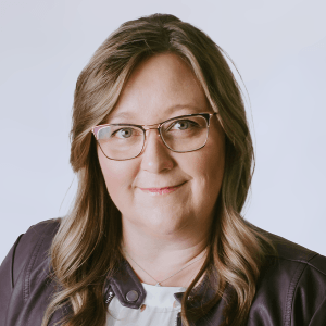 A woman with long, wavy light brown hair and glasses smiles slightly. She is wearing a dark jacket over a white top, with a light background.