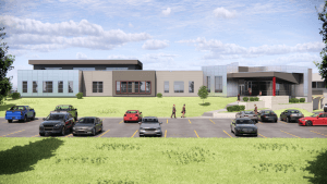 A modern building with large windows and a glass entrance, seen from a parking lot with several cars. People are walking near the entrance and across the grass under a partly cloudy sky.