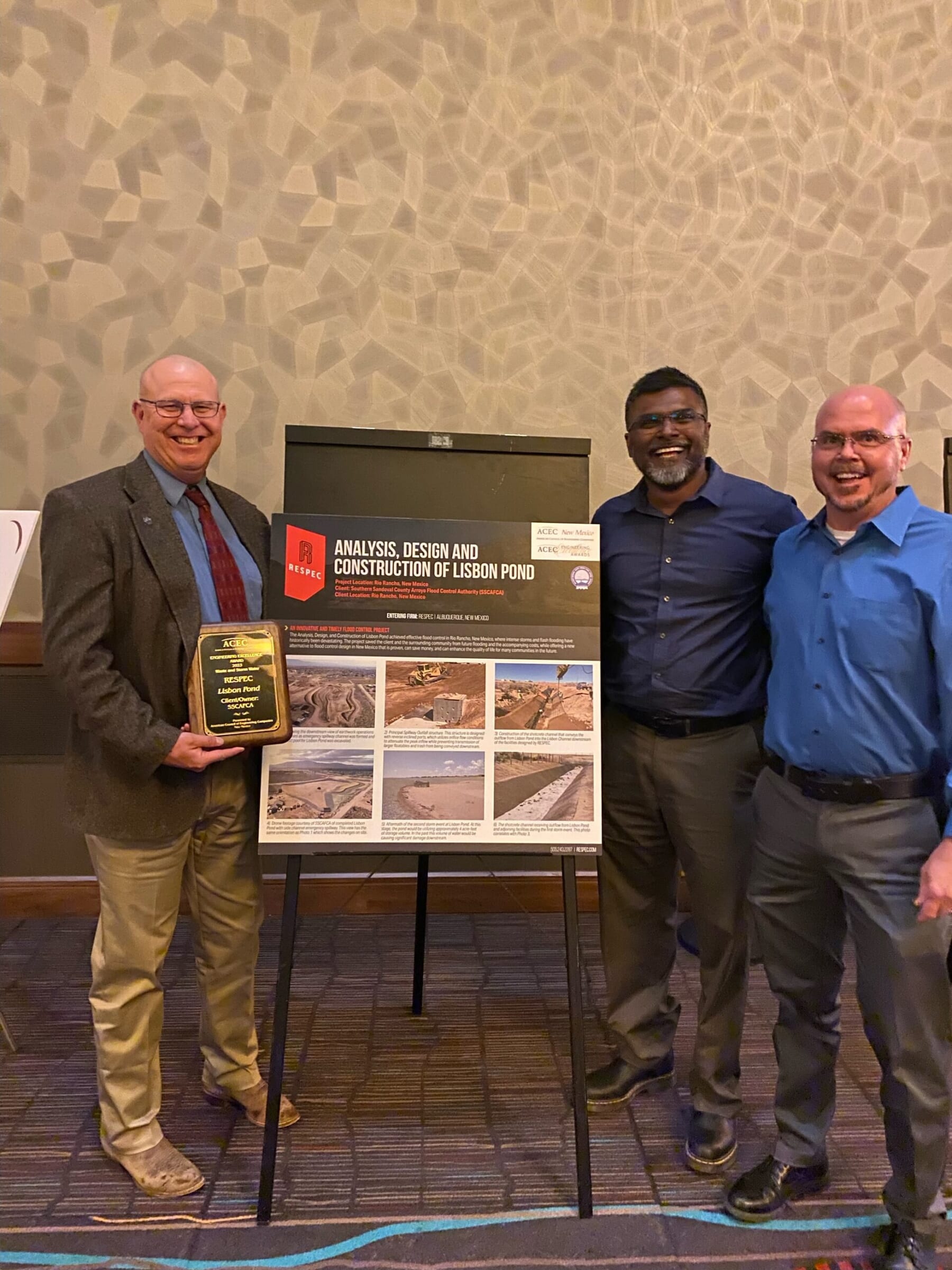 Three men stand smiling beside a display board titled “Analysis, Design and Construction of Lisbon Pond.” One man holds a plaque, and construction project photos are shown on the board behind them.