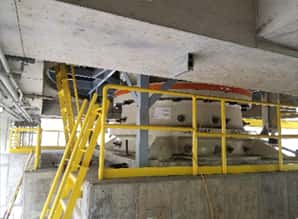 Large industrial machine with a round, white base and orange ring, surrounded by yellow safety railings and a yellow metal staircase in a concrete facility. Pipes and beams are visible overhead.