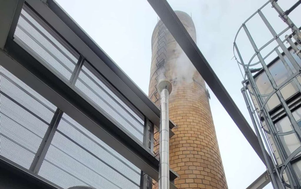A view looking up at a tall brick chimney emitting white smoke, surrounded by metal structures and safety railings against a cloudy sky.