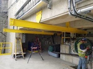 A construction worker in a safety vest and helmet stands near machinery under a concrete structure with yellow steel beams installed overhead. Ladders and tools are nearby, and electrical conduits are attached to the ceiling.