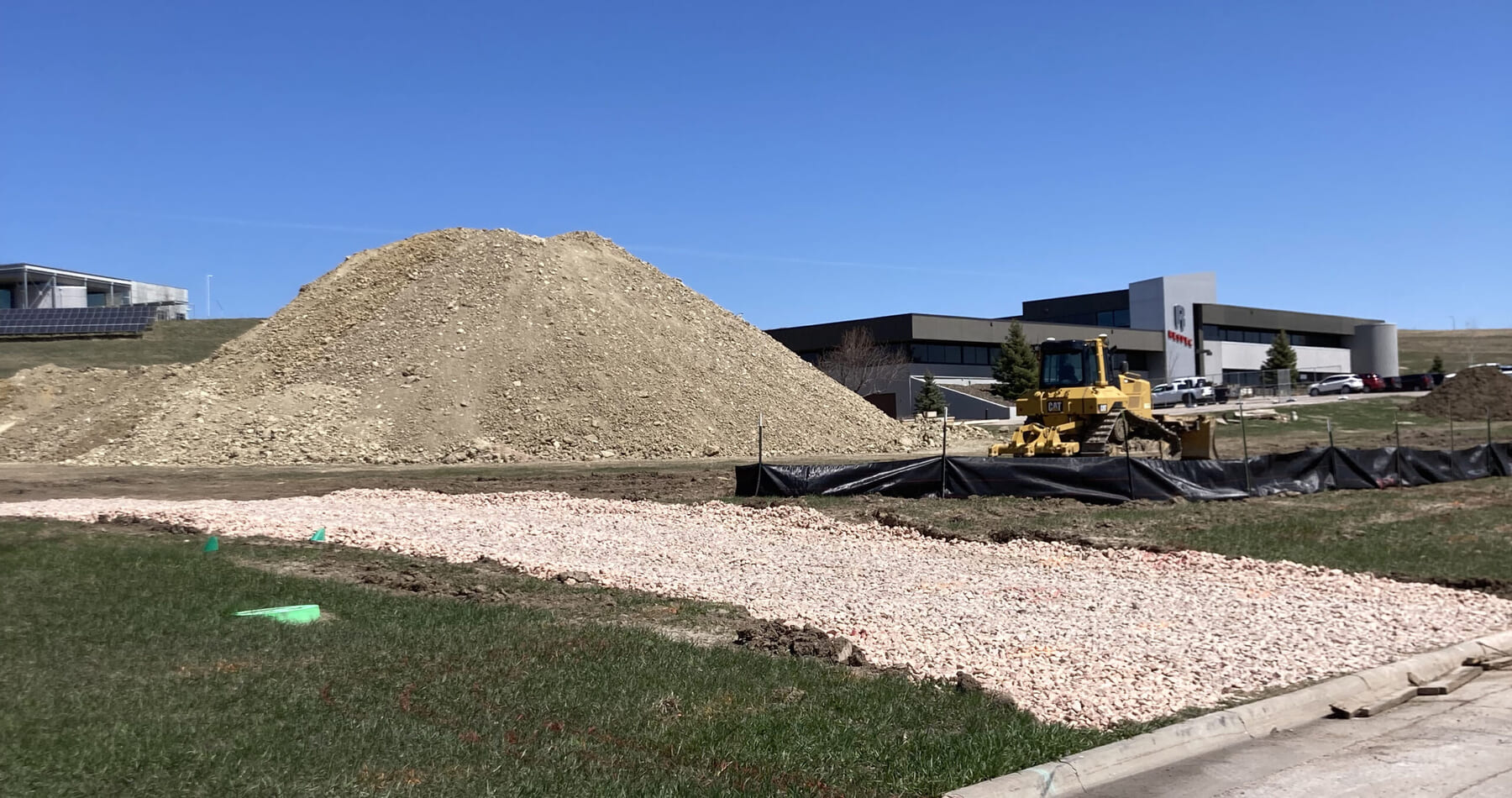 A construction site with a large dirt pile, a yellow bulldozer, gravel-covered ground, black mesh fencing, and modern buildings in the background under a clear blue sky.