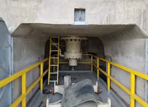 A large industrial valve is centered in a concrete enclosure with yellow safety railings and a yellow ladder on the left side. The setting appears to be part of a utility or water management facility.
