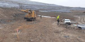 Two workers in safety gear stand near construction equipment and pickup trucks on a muddy, sloped site with large tarps covering soil in the background on an overcast day.