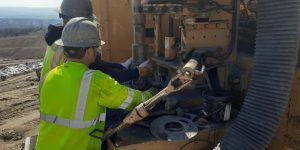 Two construction workers in safety gear operate heavy machinery at a worksite. One wears a bright yellow jacket and hard hat, while the other stands beside, both focused on the equipment in front of them.