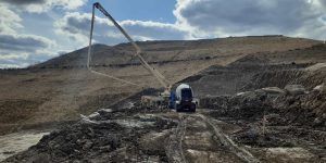 A large concrete pump truck operates on a muddy construction site near a hillside under a cloudy sky, with workers and equipment visible around the machinery.
