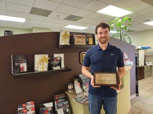 A man in a navy polo shirt stands in an office, smiling and holding a plaque that reads “Friend of Pharmacy of the Year, Joseph Stover.” Award plaques and brochures are displayed on shelves and a countertop behind him.