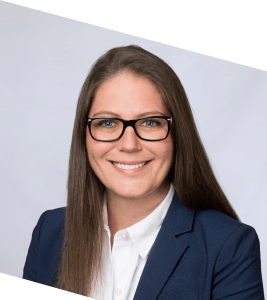 A woman with long brown hair wearing black glasses, a white shirt, and a navy blazer smiles at the camera against a plain light background.