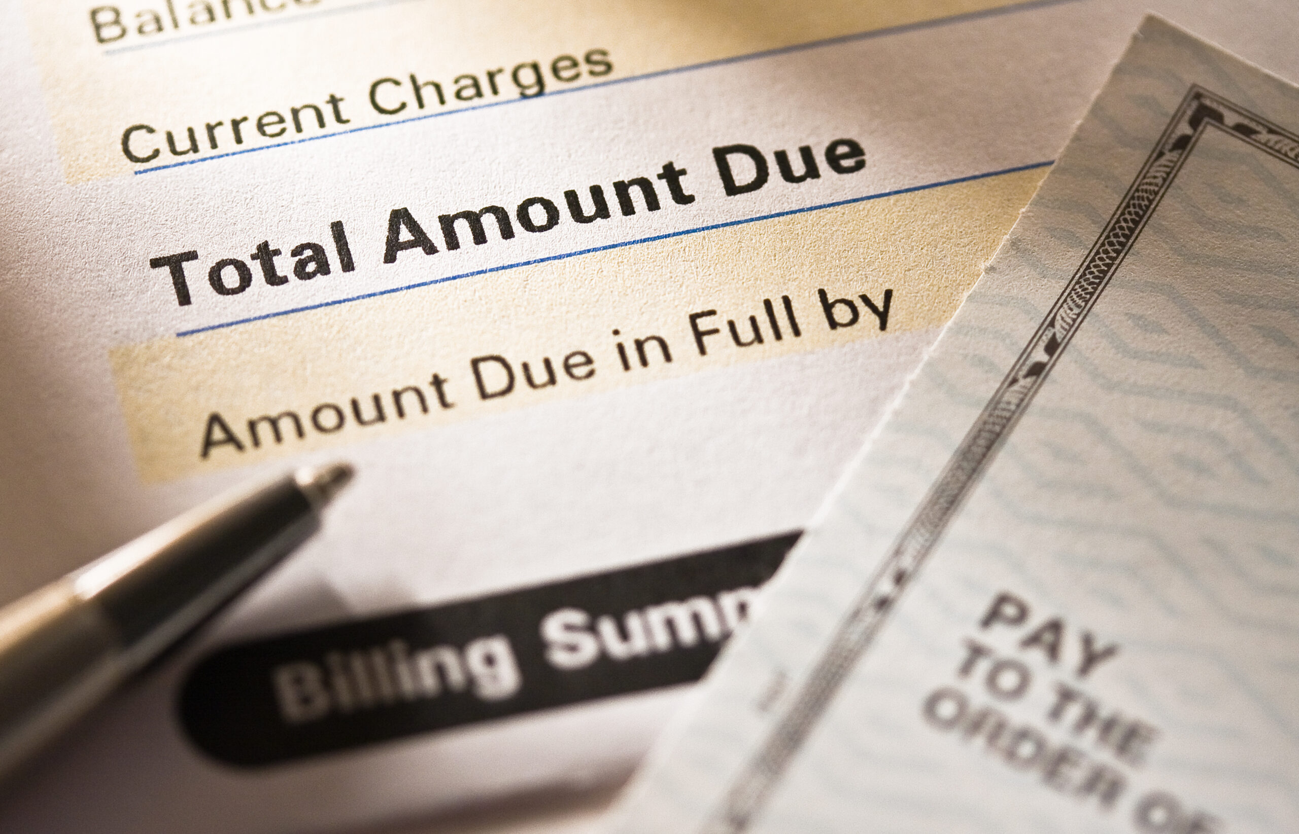 A close-up of a bill showing Total Amount Due and Amount Due in Full by next to a pen and the edge of a personal check. The scene suggests making a payment.