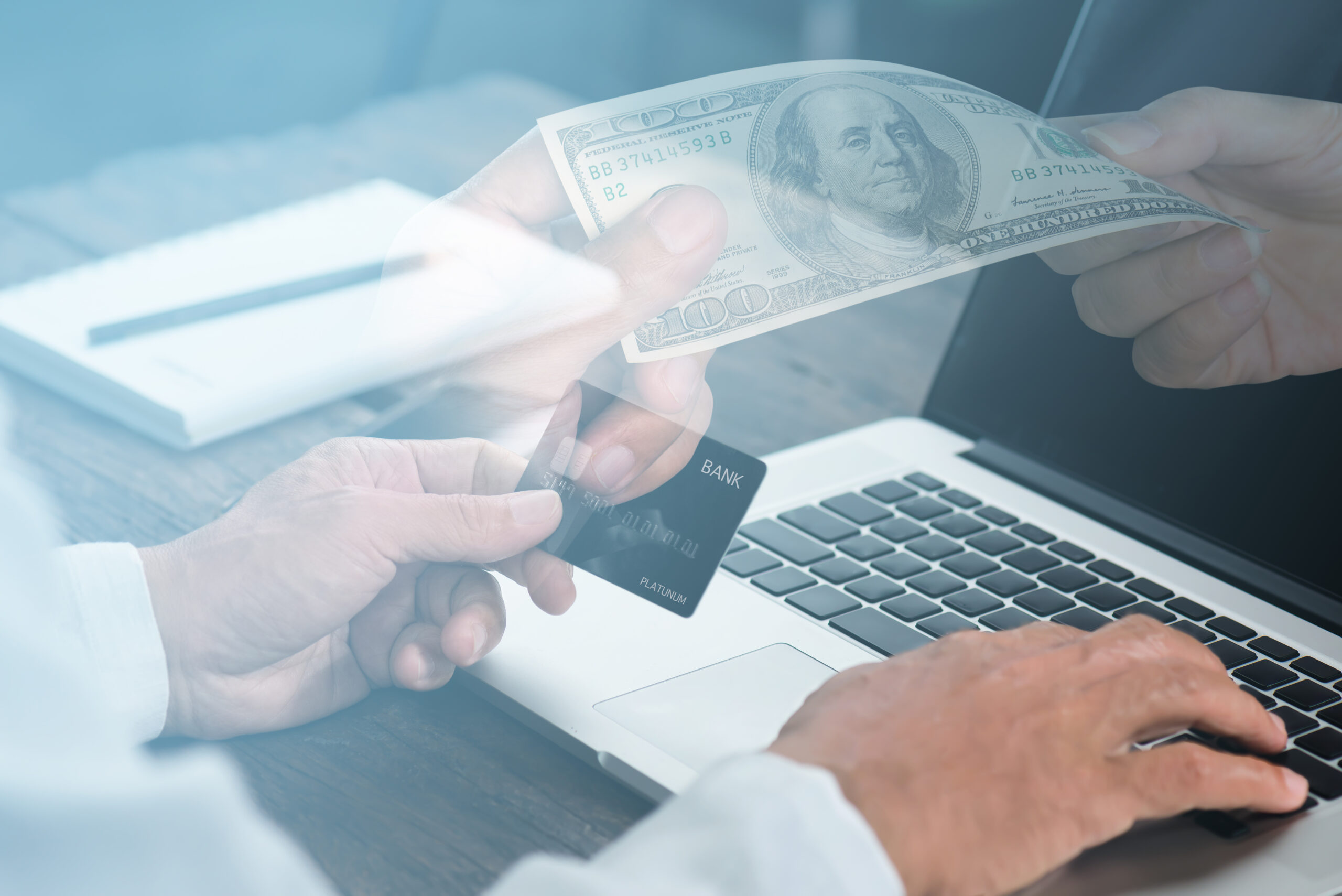 A person’s hands holding a credit card and typing on a laptop keyboard, with another hand offering a $100 bill, symbolizing online payment or financial transactions.