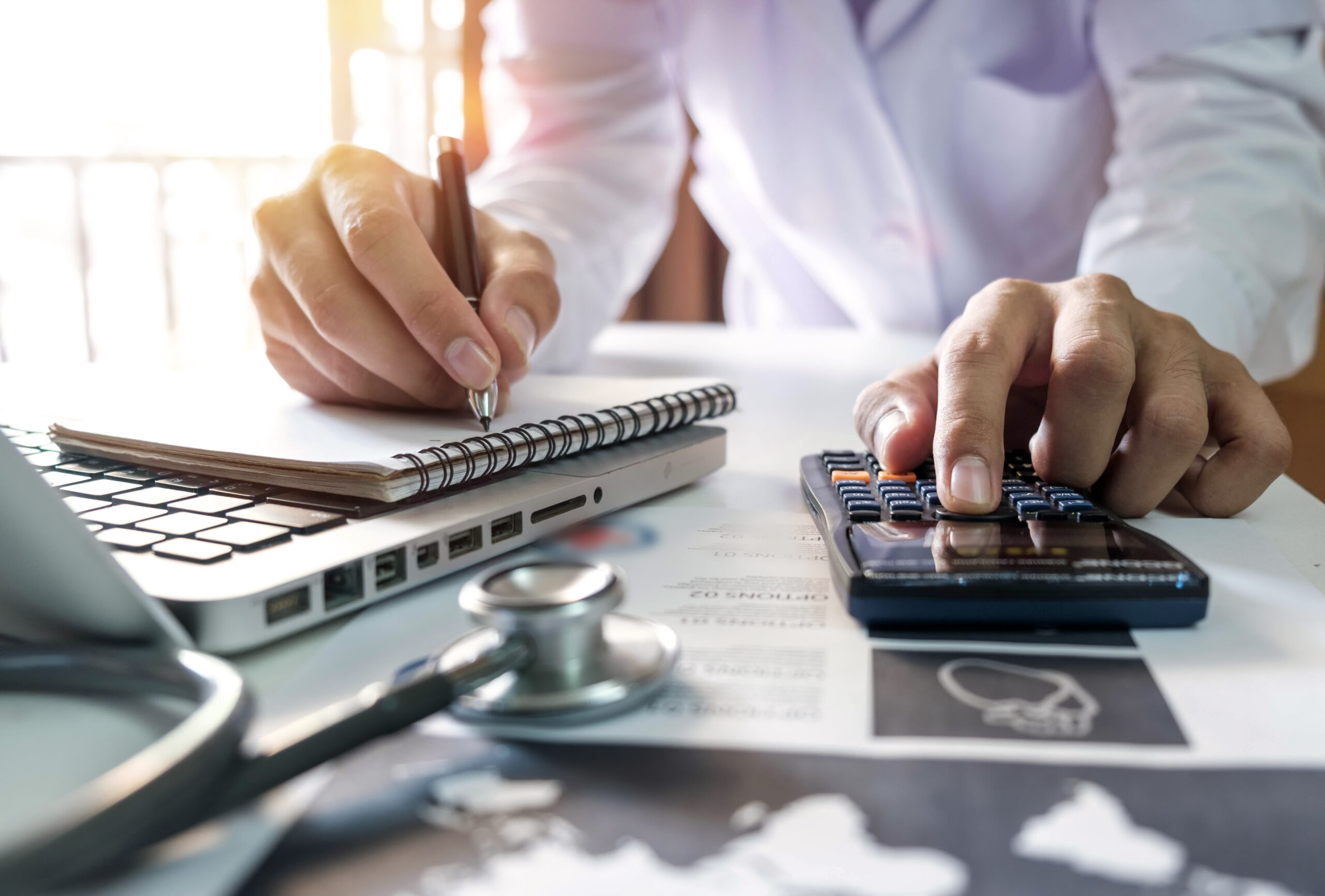 A person writes in a notebook while using a calculator, with a laptop and stethoscope on the desk, suggesting medical or healthcare financial work.