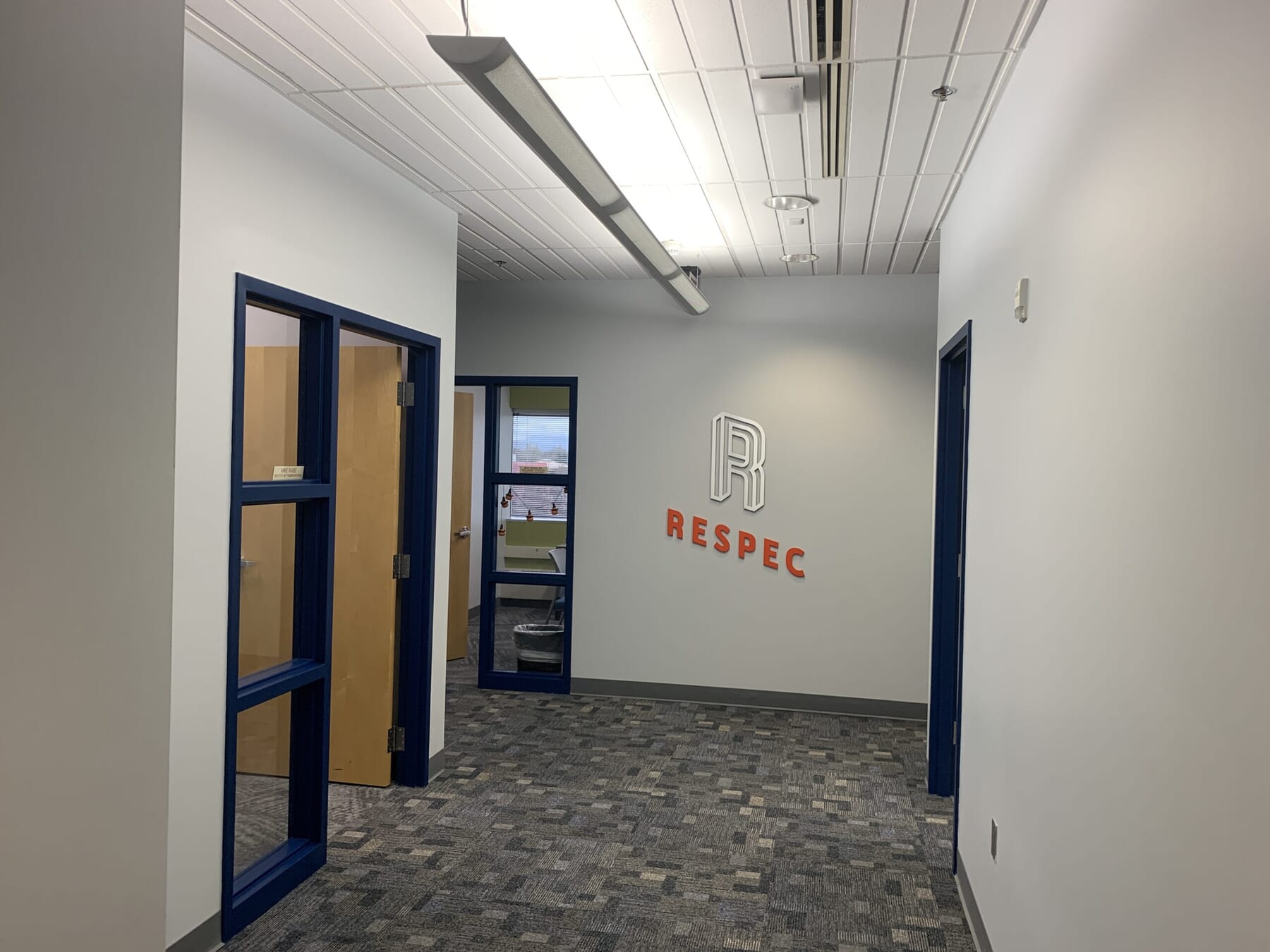 A hallway in an office building with gray carpet, white walls, and blue door frames. The word RESPEC is on the far wall in red letters, along with a stylized R logo above it. Doors lead to rooms on the left.
