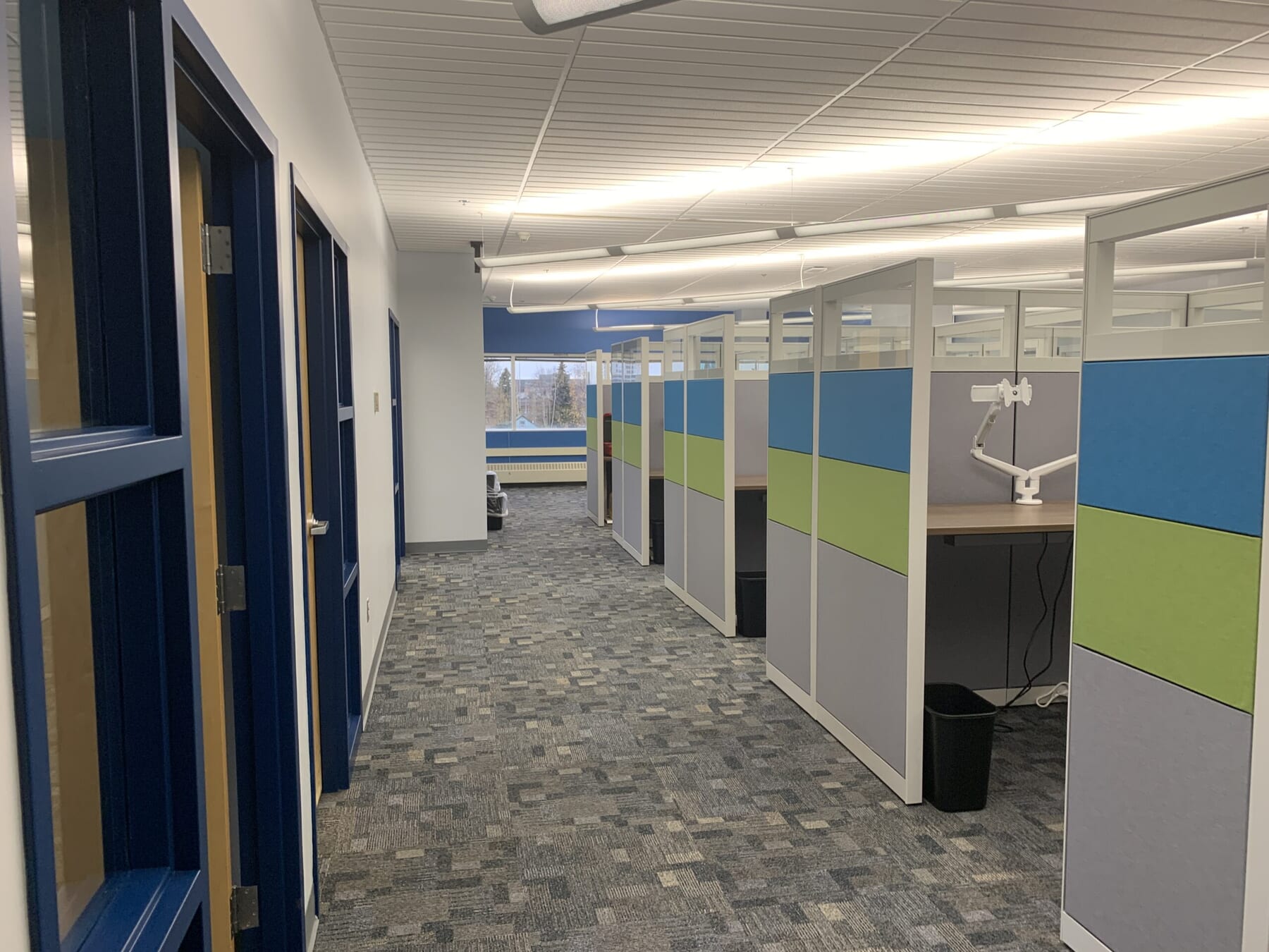 A modern office hallway with blue doors on the left and several cubicles on the right, featuring blue, gray, and green panels. The space is well-lit with large windows at the end.
