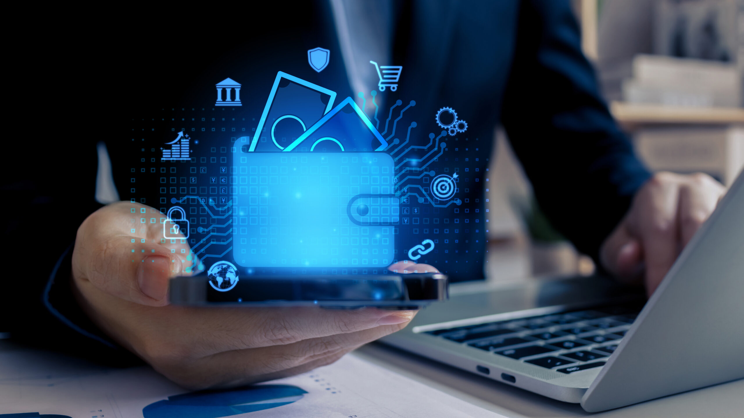 A person holds a smartphone displaying a digital wallet icon with floating money symbols and financial icons, next to an open laptop on a desk.