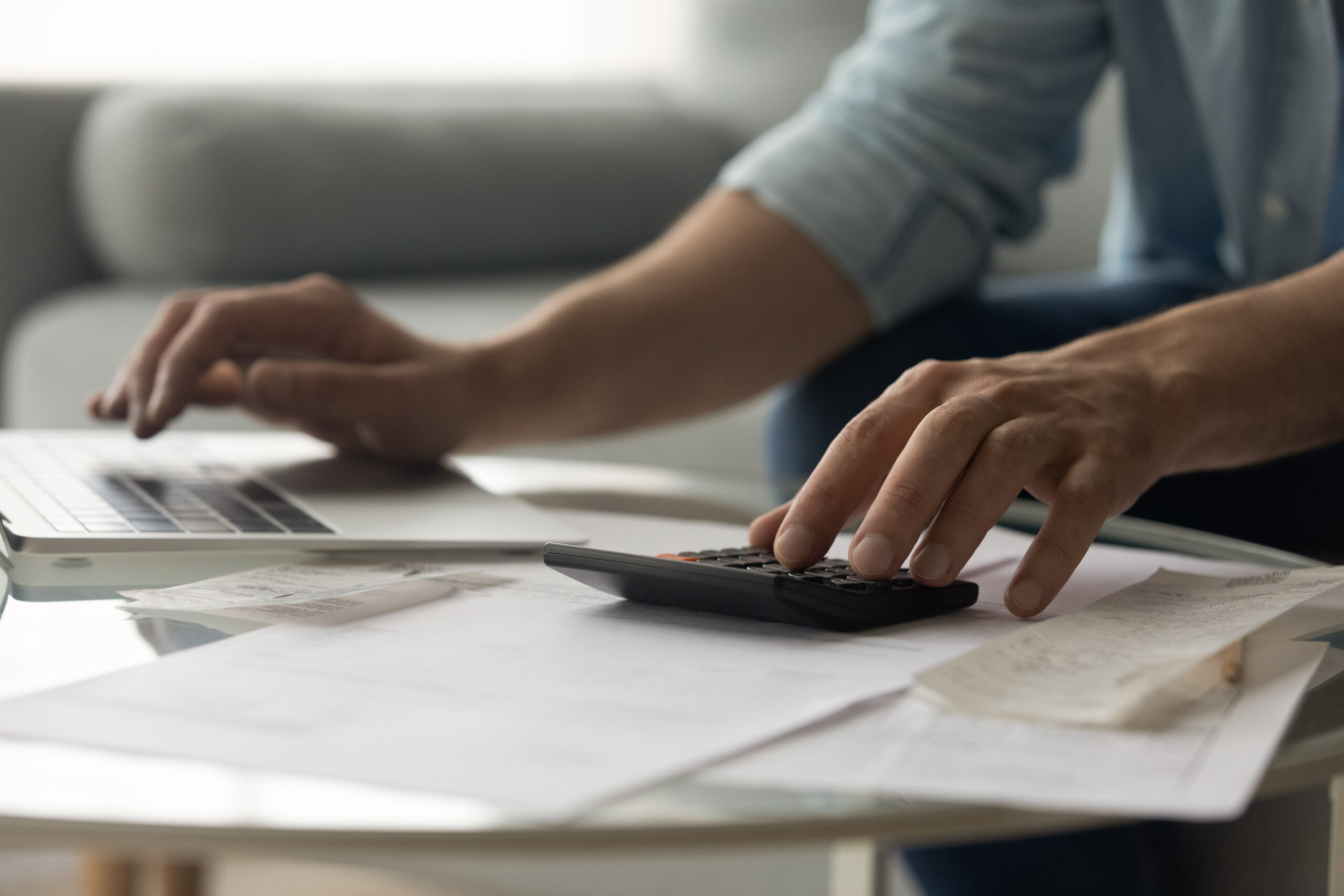 Close-up of a person’s hands using a calculator and typing on a laptop, with papers and receipts spread out on a glass table, suggesting budgeting or financial planning.