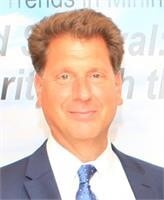 A man with short, curly brown hair wearing a dark suit, white shirt, and blue tie stands in front of a light background with blurred text. He is smiling at the camera.