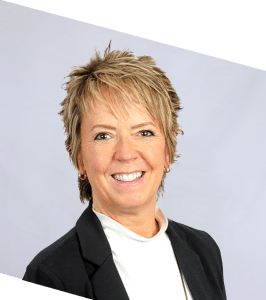A woman with short, light brown hair and a friendly smile, wearing a black blazer over a white top, poses against a plain light gray background. The image is slightly tilted.