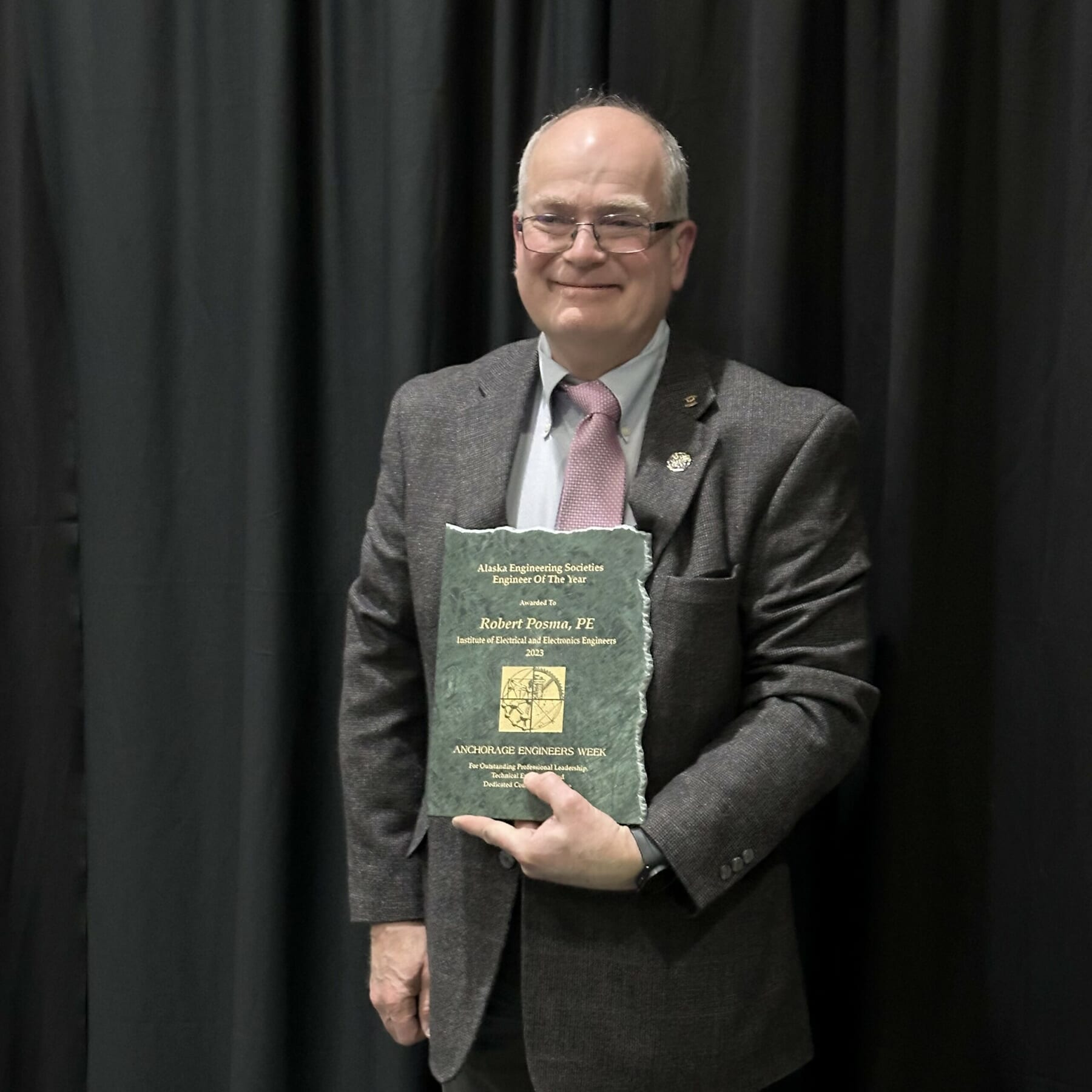 A man in a suit and glasses stands in front of a dark curtain, smiling and holding a plaque that reads “Alaska Engineering Societies Engineer of the Year” and “Robert E. Brown, PE” along with other text.