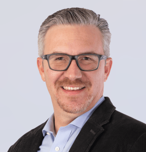 A smiling man with short gray hair, glasses, and a goatee, wearing a light blue collared shirt and a dark blazer, poses against a light background.