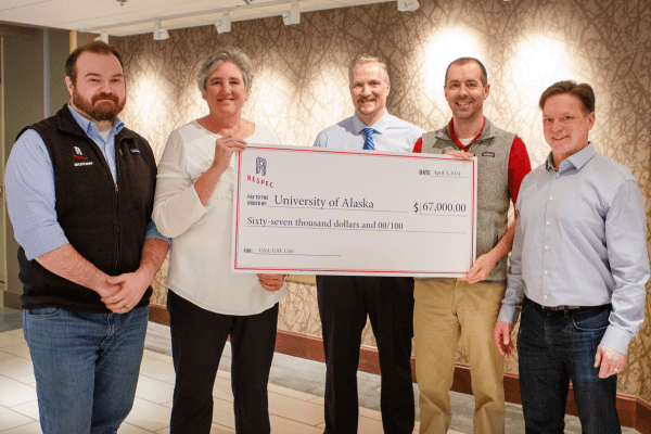 Five people stand indoors, smiling and holding a large ceremonial check for $67,000 made out to the University of Alaska from RESPEC. The group is dressed in business casual attire.