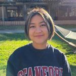 A person with short hair and a dark Stanford sweatshirt smiles outdoors in a sunny grassy area, with a hammock and buildings visible in the background.