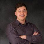 A young man with short brown hair smiles at the camera, standing with arms crossed. He is wearing a dark button-up shirt against a dark, plain background.