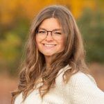A young woman with long wavy brown hair and glasses smiles outdoors, wearing a white sweater. The background is blurred with warm autumn colors of yellow, orange, and green.
