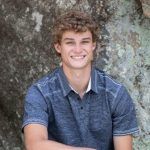 A young man with curly brown hair, wearing a short-sleeved blue button-up shirt, sits and smiles in front of a large, textured rock surface.