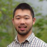 A young man with short black hair and a trimmed beard is smiling at the camera. He is wearing a plaid shirt and standing outdoors with green foliage in the background.