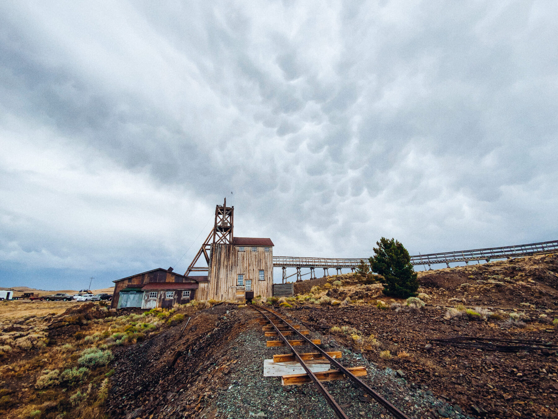 An old wooden mining structure sits on rocky terrain under a cloudy, textured sky, with rusty railroad tracks leading up to it and a conveyor belt stretching across the landscape.