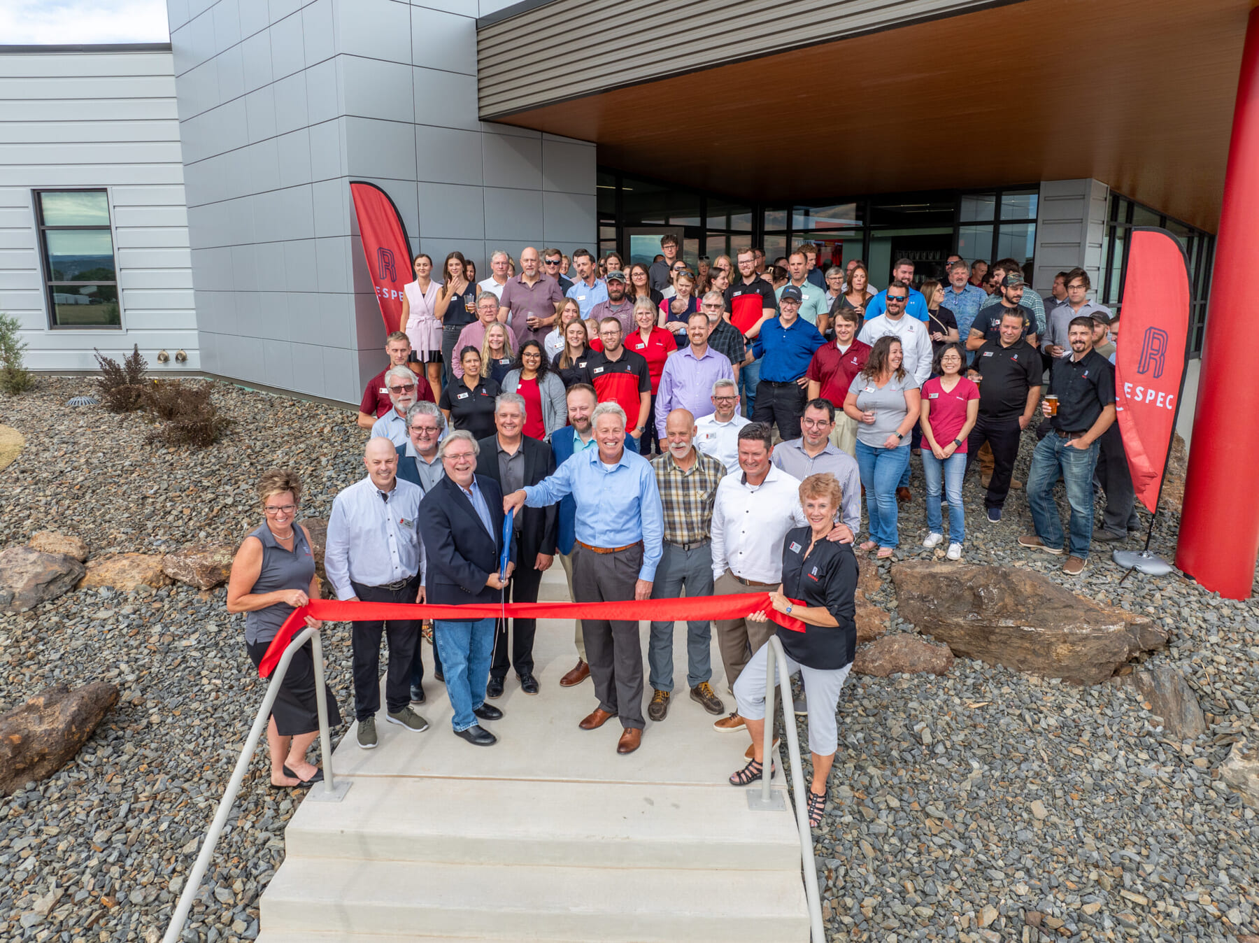 A large group of people stands outside a modern building, smiling and posing for a photo during a ribbon-cutting ceremony. Several people in front hold scissors and a red ribbon on the steps leading to the entrance.