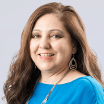 Smiling woman with long brown hair wearing a blue top, large teardrop earrings, and a necklace, posing in front of a plain light gray background.