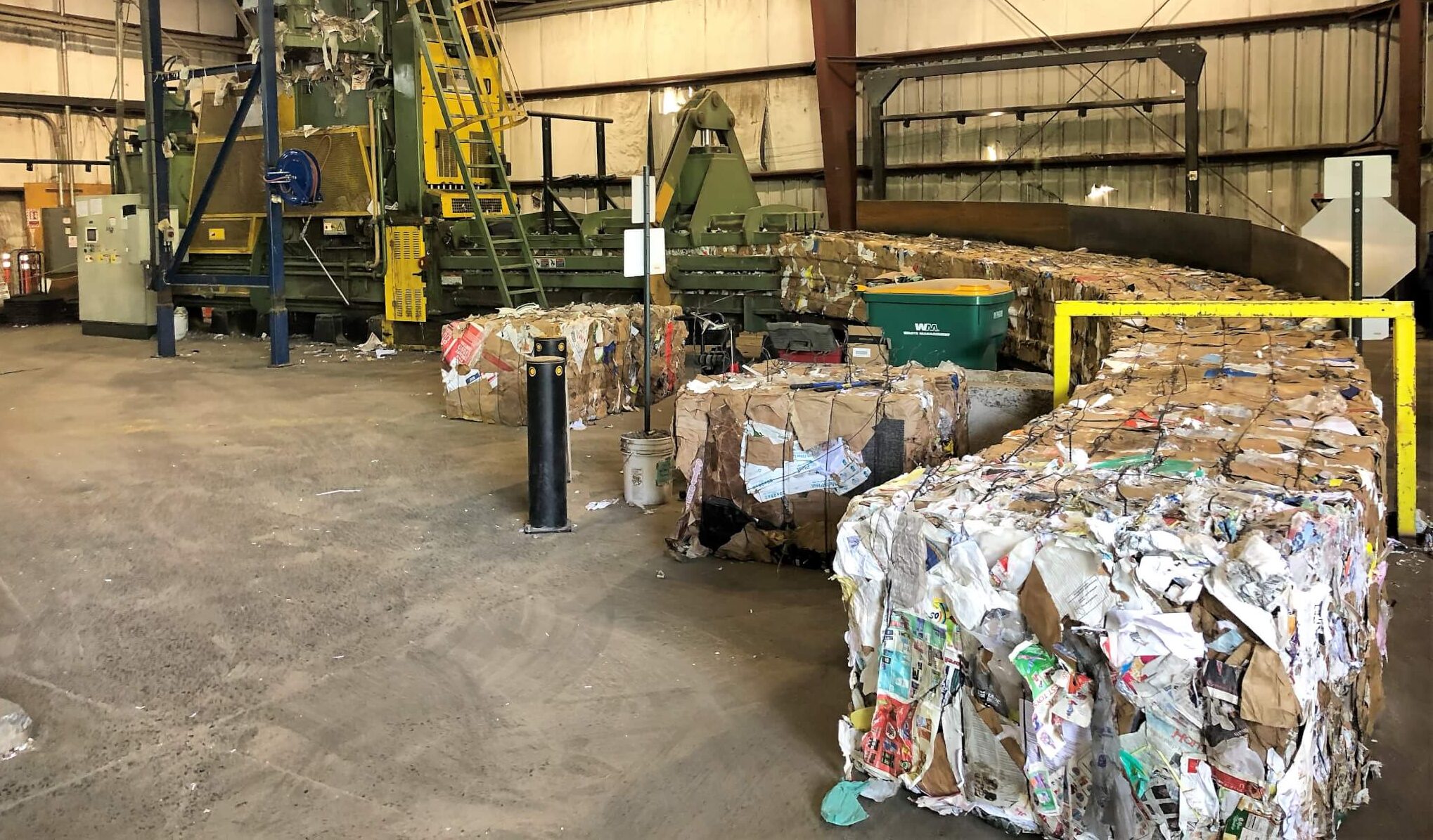 Large bales of compressed cardboard and paper are stacked in a recycling facility, with machinery and conveyor belts in the background, and a concrete floor covered in dust and debris.