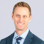 A man with short light brown hair wearing a blue suit, striped tie, and checkered shirt is smiling at the camera against a light gray background.
