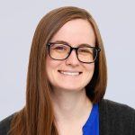 A woman with long brown hair and glasses smiles at the camera. She is wearing a blue top and a dark cardigan, in front of a light gray background.