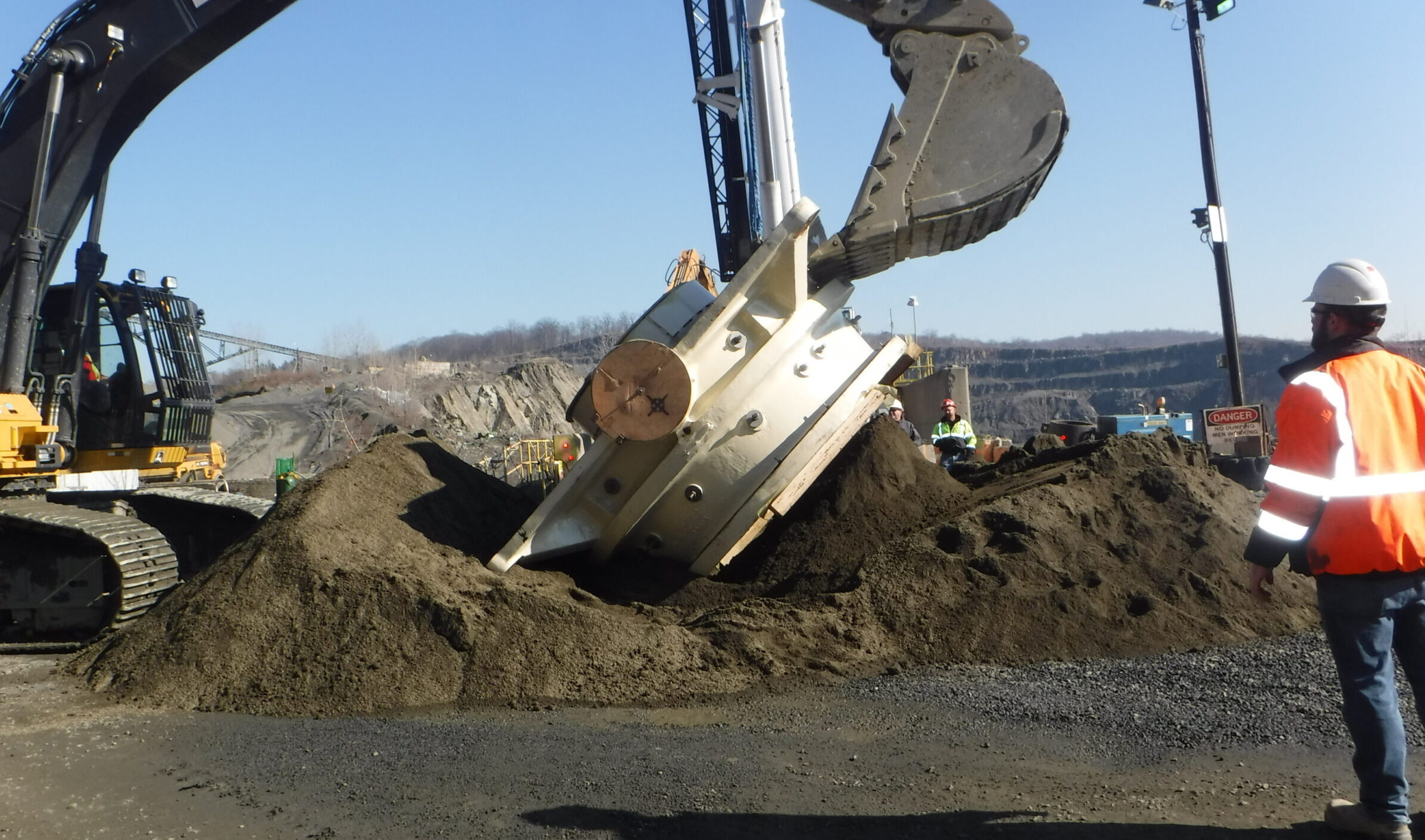 A construction site where a large excavator is lifting a cylindrical metal object partially buried in dirt, with workers in safety gear observing nearby.