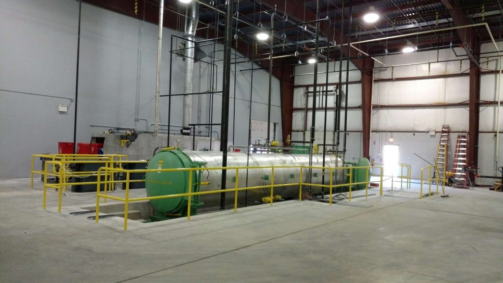 Large industrial facility interior with concrete floors, yellow safety railings, a horizontal green cylindrical tank, red barrels, and visible pipes and ladders. Bright lighting and high ceiling with exposed beams.